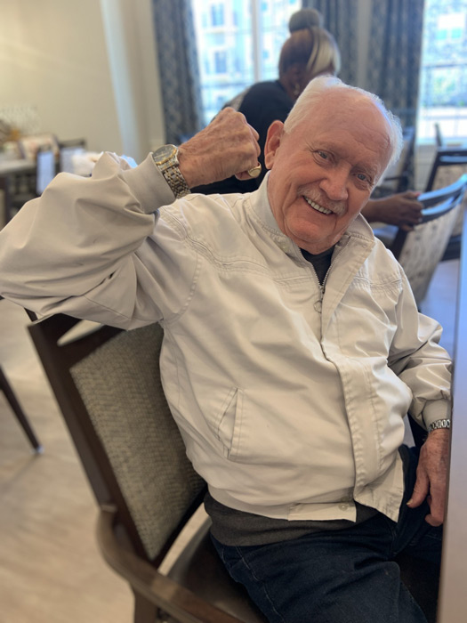 Smiling elderly man wearing a white jacket, sitting on a chair and playfully flexing his arm in a dining area.