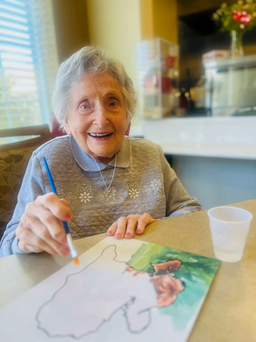 Elderly woman smiling and painting a picture at a table.