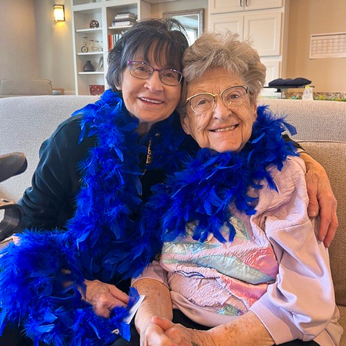 Two senior women, both wearing glasses and bright blue feather boas, sitting close together on a couch, smiling and happy to see each other in a senior living community.