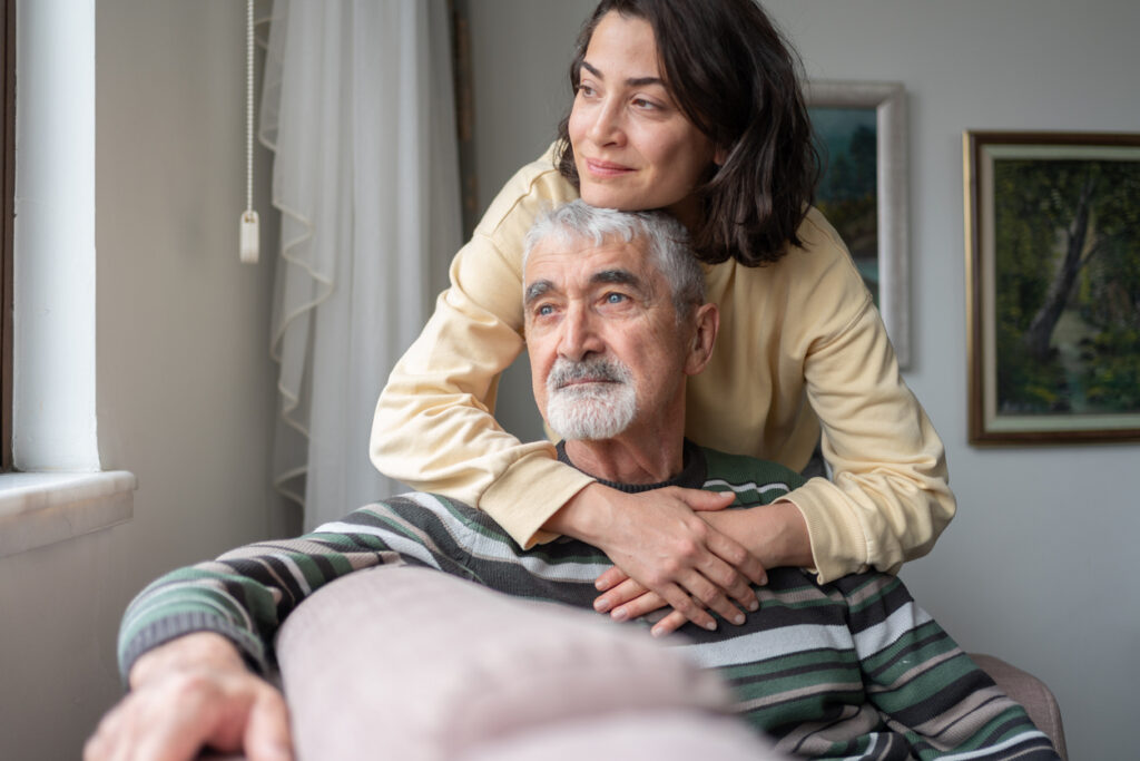 Adult daughter Hugging seated Senior Father from behind, her arms around his shoulders.