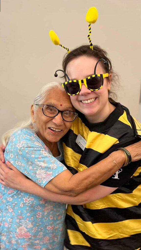 A woman in a bee costume hugs a senior woman in a blue shirt with pink and white flowers.