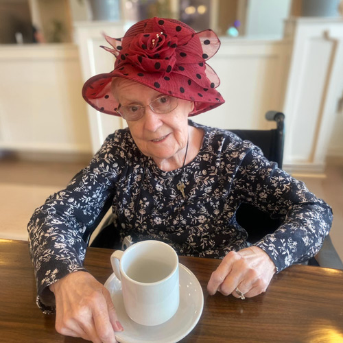 Elderly woman wearing a red hat with black polka dots and a floral dress, sitting at a table with a white cup and saucer at a senior living community.