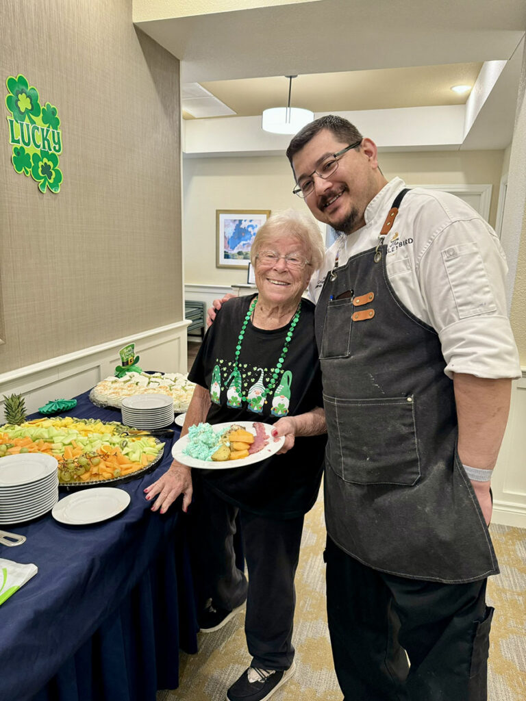 A senior woman stands by a St. Patrick's Day spread holding a plate of food. A chef stands beside her with his arm around her shoulder.