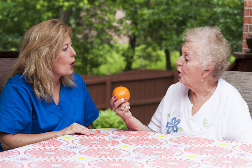 Middle-aged female speech therapist with a senior woman sitting at a table outdoors. The senior is holding an orange and practicing the production of a sound.