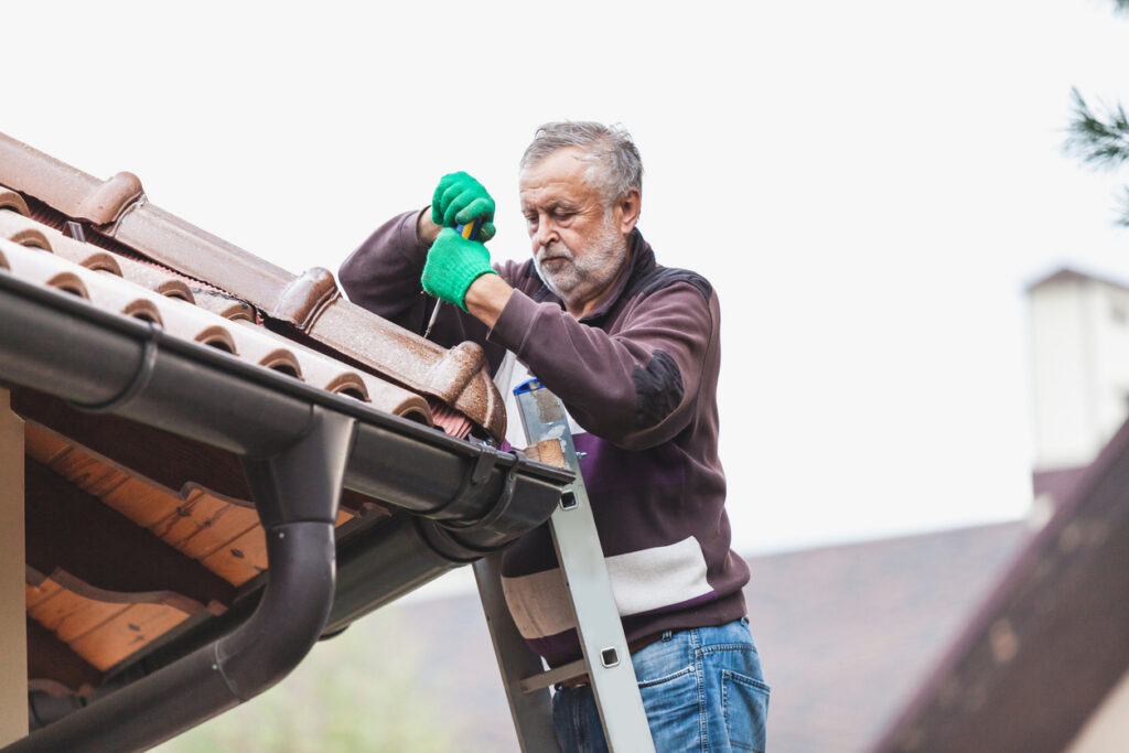 senior man with a gray beard stands on a metal ladder and repairs a ceramic shingle roof