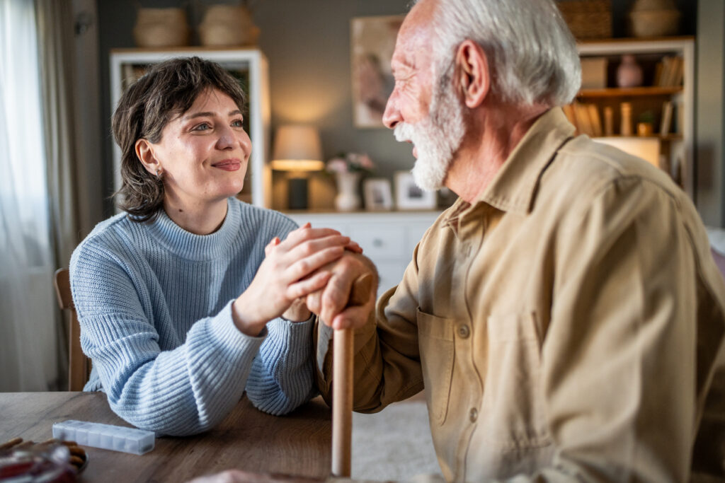 Young woman smiling while holding hands with a senior man, who uses a cane, sharing a warm connection in a cozy room during the daytime