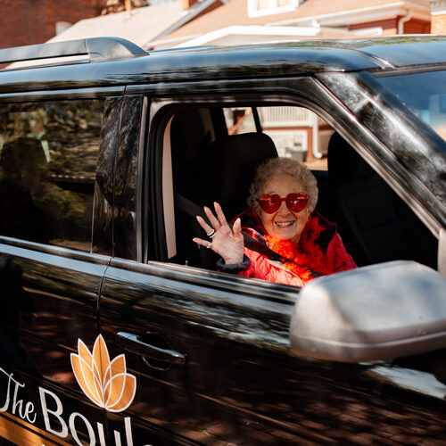 senior woman in the passenger seat of a senior living community vehicle, smiling and waving to the camera